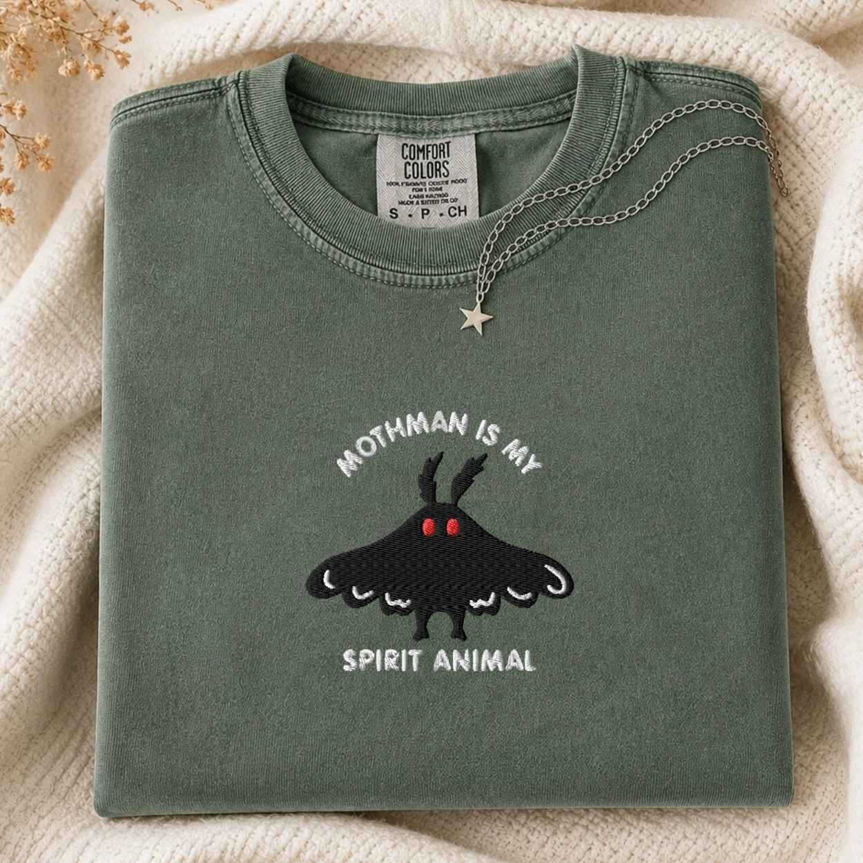 A folded sage green t-shirt with a black moth graphic and the text 'MOTHMAN IS MY SPIRIT ANIMAL' on the front, styled on a cream knit blanket with a silver necklace.