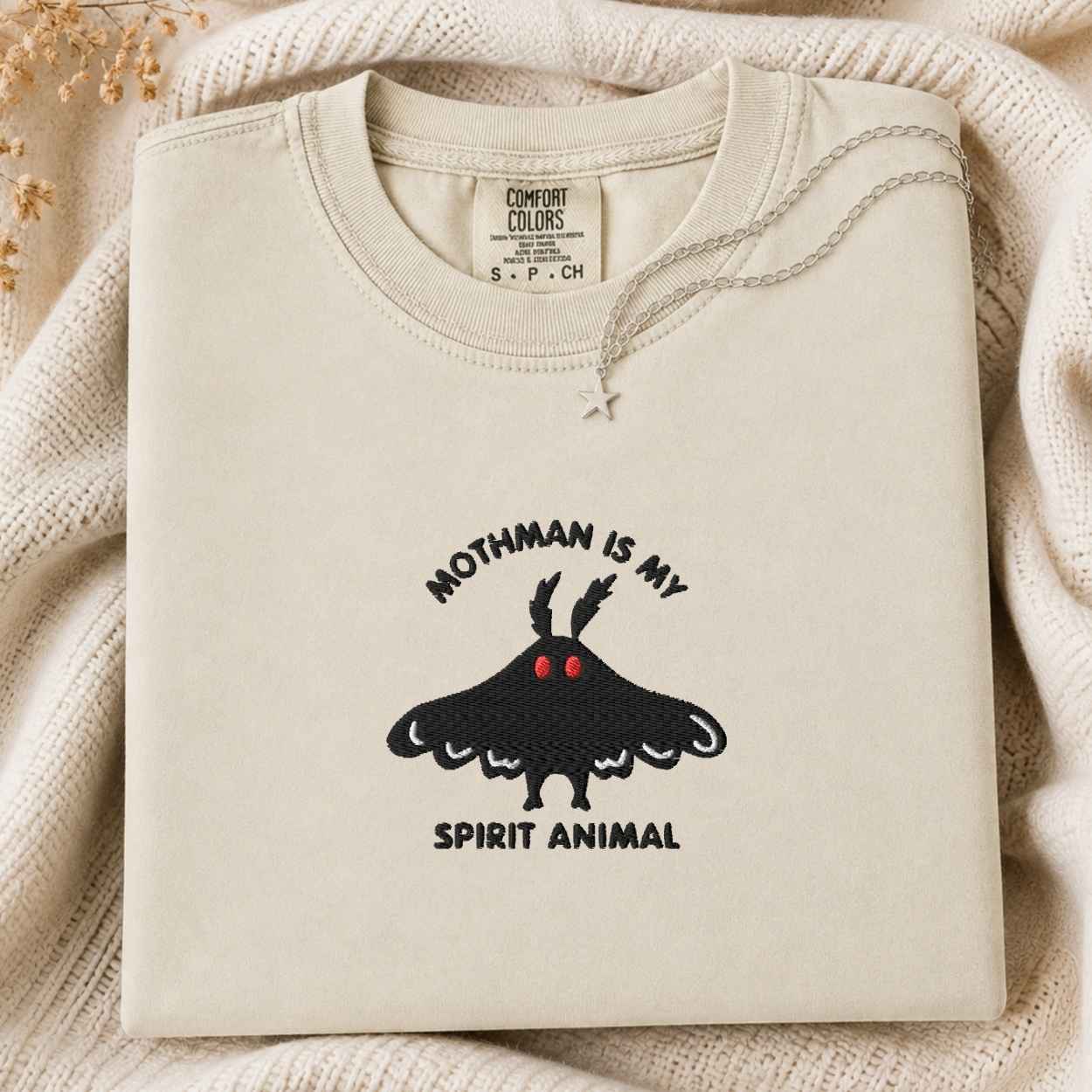 A folded cream-colored tshirt with a black moth graphic and the text 'MOTHMAN IS MY SPIRIT ANIMAL' on the front, styled on a textured cream knit blanket with dried flowers in the corner.