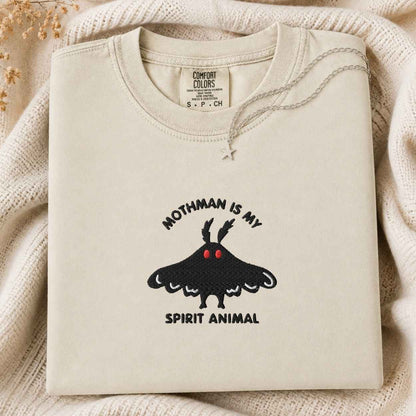 A folded cream-colored tshirt with a black moth graphic and the text 'MOTHMAN IS MY SPIRIT ANIMAL' on the front, styled on a textured cream knit blanket with dried flowers in the corner.