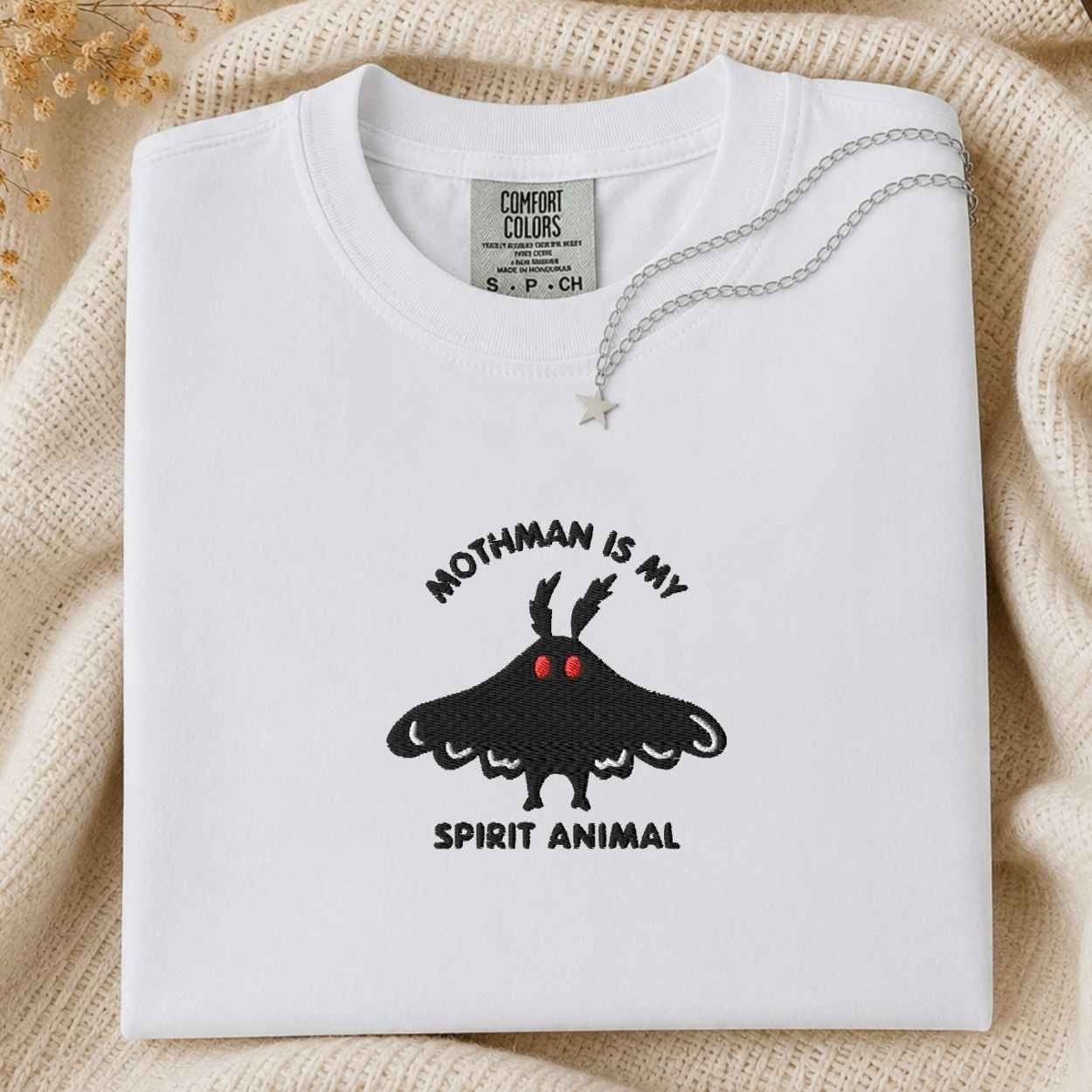 A folded white Comfort Colors t-shirt with a black embroidered moth graphic and the text 'MOTHMAN IS MY SPIRIT ANIMAL' in black and red. The shirt is styled on a beige knit blanket with a silver necklace draped over the collar.