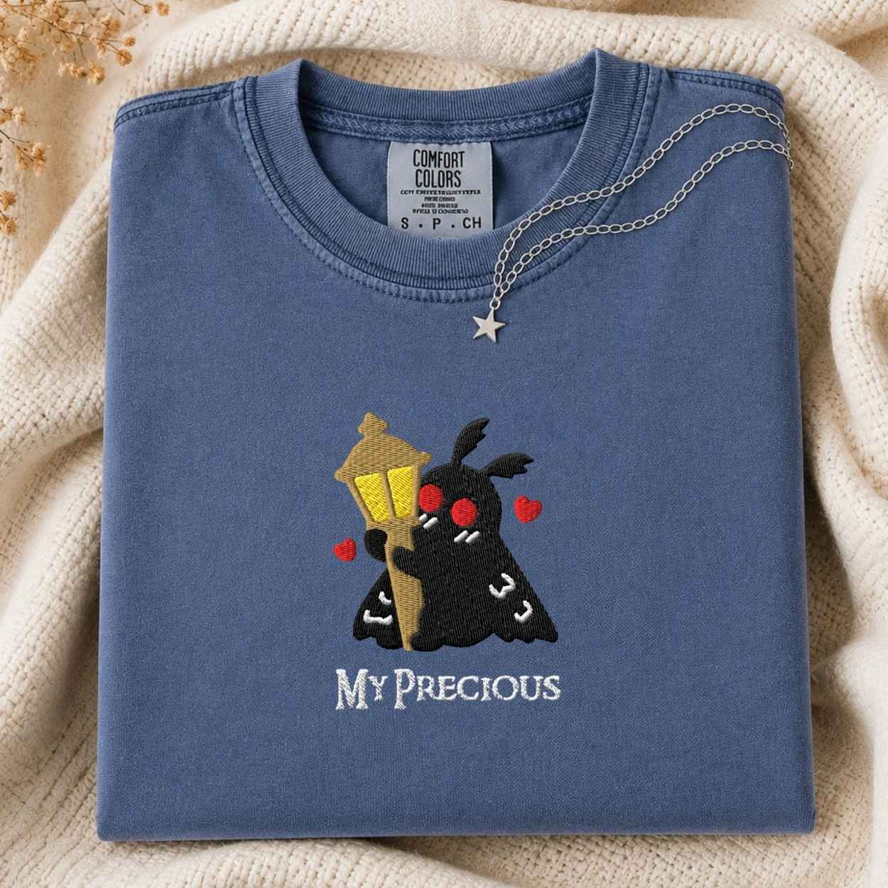 A folded slate blue t-shirt with a custom embroidered design of a black moth holding a golden street lamp, surrounded by red hearts and the text 'MY PRECIOUS' in white. A silver star necklace is draped over the collar.