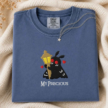 A folded slate blue t-shirt with a custom embroidered design of a black moth holding a golden street lamp, surrounded by red hearts and the text 'MY PRECIOUS' in white. A silver star necklace is draped over the collar.