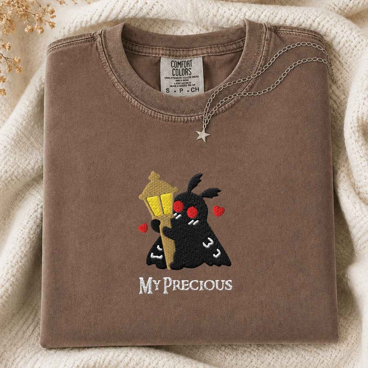 A folded brown tshirt with a cute embroidered graphic of a black moth holding a yellow street lamp and wearing red glasses, with the text 'MY PRECIOUS' below it. The tshirt is styled on a cream-colored knit blanket with a silver star necklace draped over the collar.