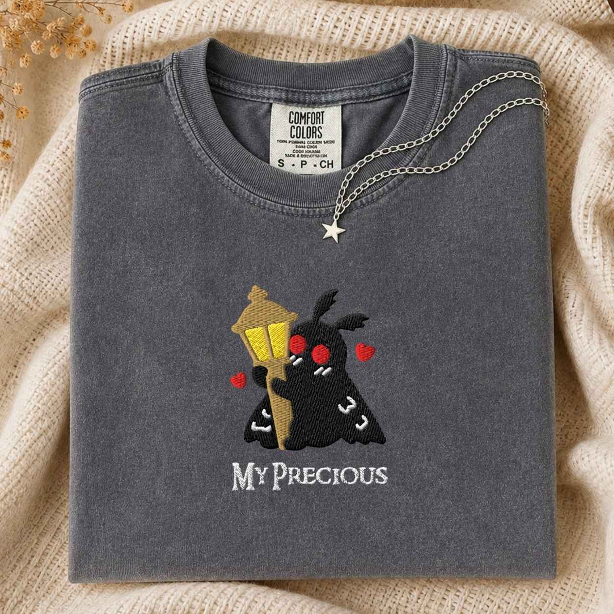 A folded dark gray Comfort Colors t-shirt with a custom embroidered design of a black mothman holding a yellow street lamp, red hearts, and the text 'MY PRECIOUS' in white. The shirt is styled on a beige knit blanket with a silver star necklace draped over the collar.