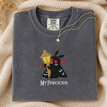 A folded dark gray Comfort Colors t-shirt with a custom embroidered design of a black mothman holding a yellow street lamp, red hearts, and the text 'MY PRECIOUS' in white. The shirt is styled on a beige knit blanket with a silver star necklace draped over the collar.