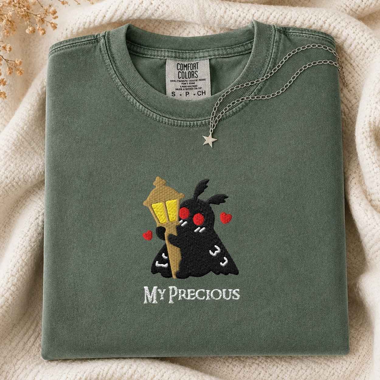 A folded sage green t-shirt with a cute embroidered design of a black moth holding a yellow street lamp and red hearts, with the text 'MY PRECIOUS' below. The shirt is styled on a cream knit blanket with a silver star necklace draped over it.