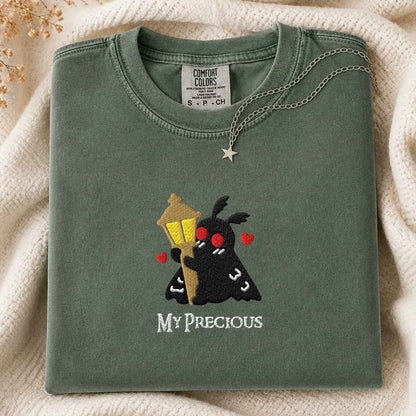 A folded sage green t-shirt with a cute embroidered design of a black moth holding a yellow street lamp and red hearts, with the text 'MY PRECIOUS' below. The shirt is styled on a cream knit blanket with a silver star necklace draped over it.