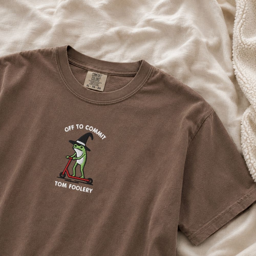 A brown short-sleeve t-shirt featuring a graphic of a green cartoon frog wearing a witch hat and riding a red scooter, with the text 'OFF TO COMMIT TOM FOOLERY'. The shirt is laid flat on a beige fabric background.