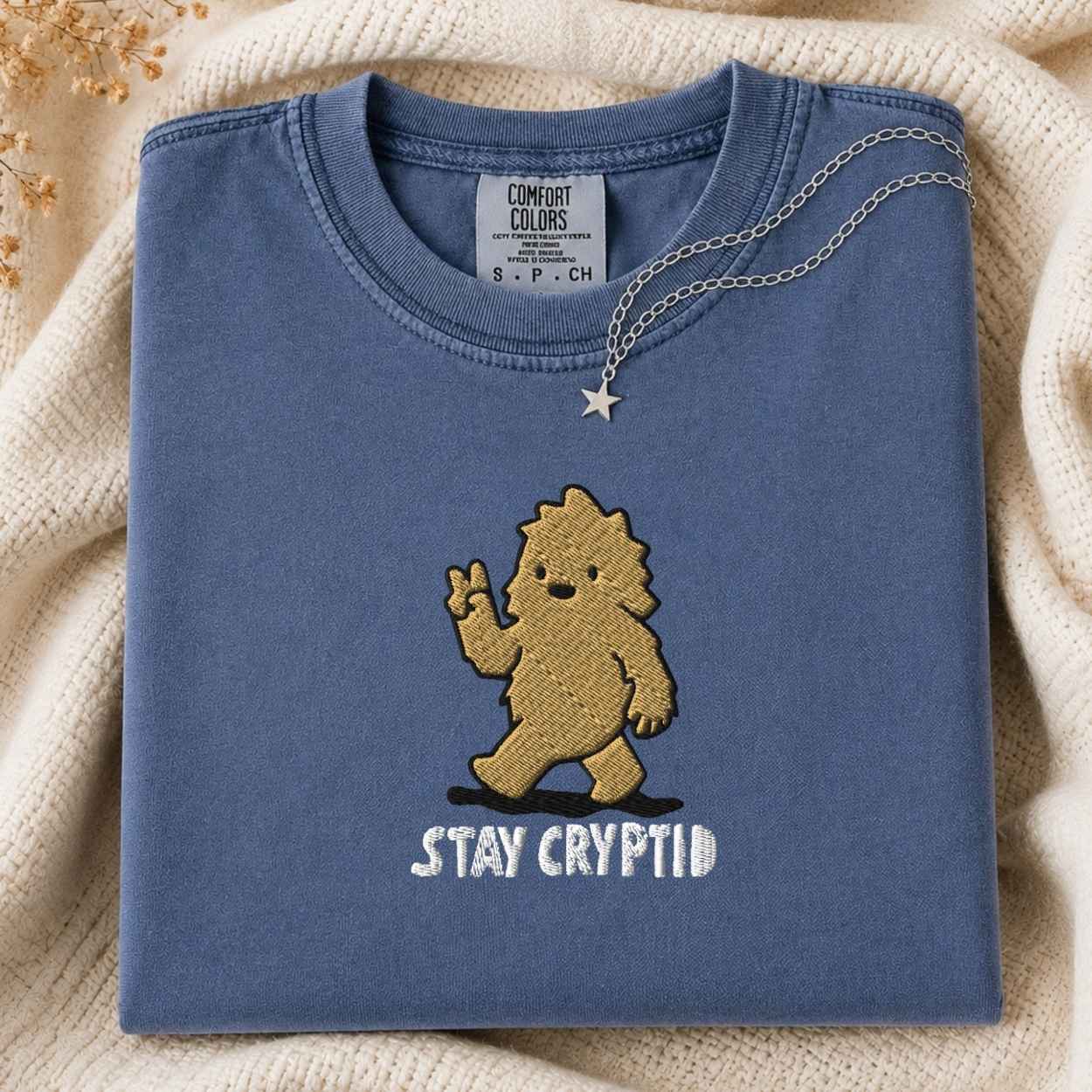 A folded slate blue t-shirt with a  embroidered cartoon bigfoot and the white embroidered text 'STAY CRYPTID' on the front, styled on a cream knit blanket with a silver necklace draped over the collar.