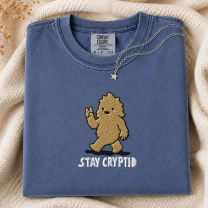 A folded slate blue t-shirt with a  embroidered cartoon bigfoot and the white embroidered text 'STAY CRYPTID' on the front, styled on a cream knit blanket with a silver necklace draped over the collar.