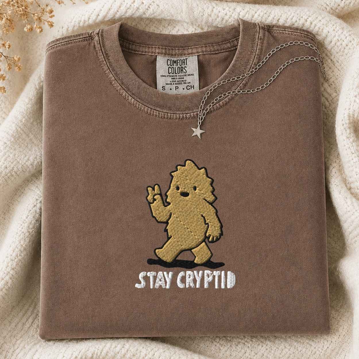 A folded brown tshirt with a gold embroidered cartoon bigfoot and the text 'STAY CRYPTID' in white, styled on a cream knit blanket with a silver necklace draped over the collar. The neck label reads 'COMFORT COLORS'.