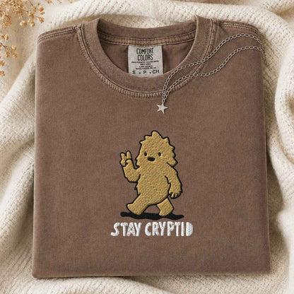 A folded brown tshirt with a gold embroidered cartoon bigfoot and the text 'STAY CRYPTID' in white, styled on a cream knit blanket with a silver necklace draped over the collar. The neck label reads 'COMFORT COLORS'.