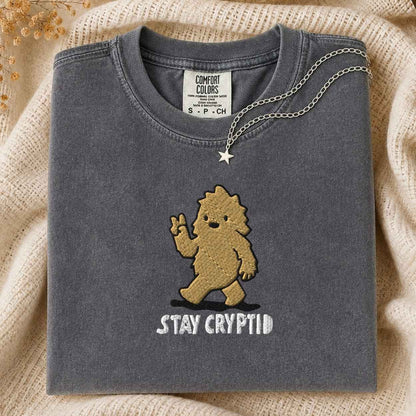 A folded dark gray t-shirt with a  embroidered cartoon bigfoot and the text 'STAY CRYPTID' in white, styled on a beige knit blanket with a silver star necklace draped over the collar.