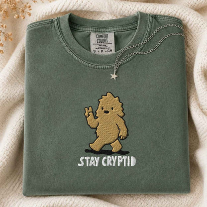 A folded sage green Comfort Colors t-shirt featuring an embroidered cartoon creature and the white text 'STAY CRYPTID', styled on a cream knit blanket with a silver necklace.