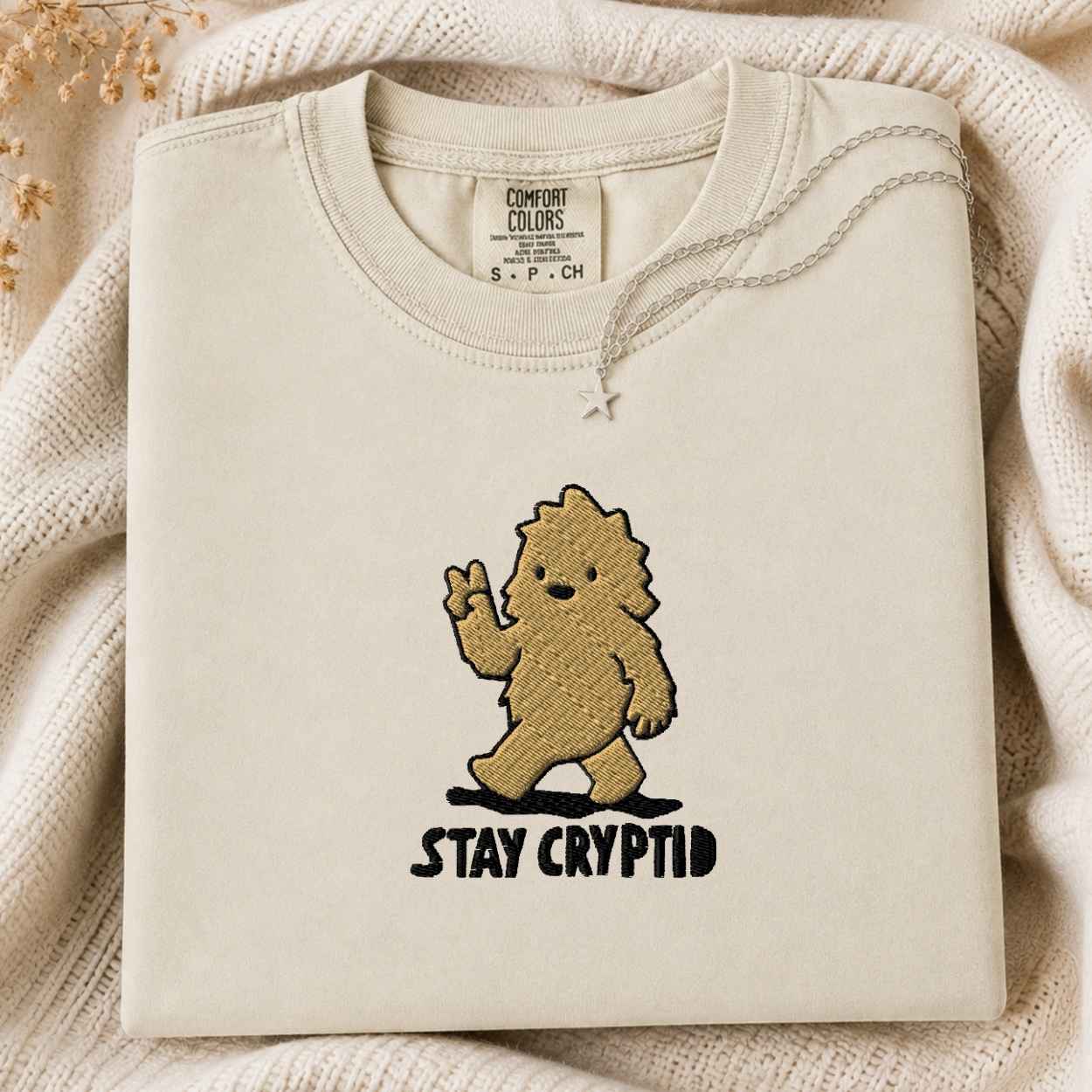 A folded cream-colored tshirt with a graphic of a golden-brown furry creature waving and the text 'STAY CRYPTID' underneath, styled on a cozy cream knit blanket.