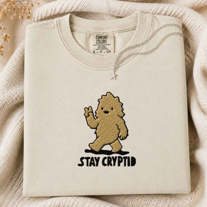 A folded cream-colored tshirt with a graphic of a golden-brown furry creature waving and the text 'STAY CRYPTID' underneath, styled on a cozy cream knit blanket.