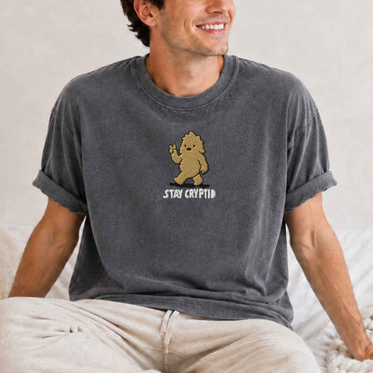 A man wearing a dark gray graphic t-shirt with a cartoon bigfoot and the text 'stay cryptid'.