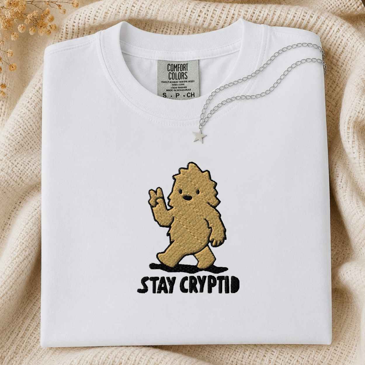 A folded white Comfort Colors t-shirt with a brown embroidered cartoon creature and the text 'STAY CRYPTID' in black. A silver necklace with a star pendant is draped over the collar.
