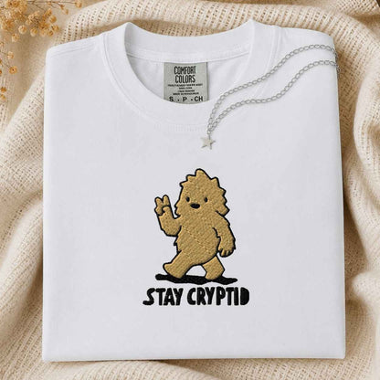 A folded white Comfort Colors t-shirt with a brown embroidered cartoon creature and the text 'STAY CRYPTID' in black. A silver necklace with a star pendant is draped over the collar.