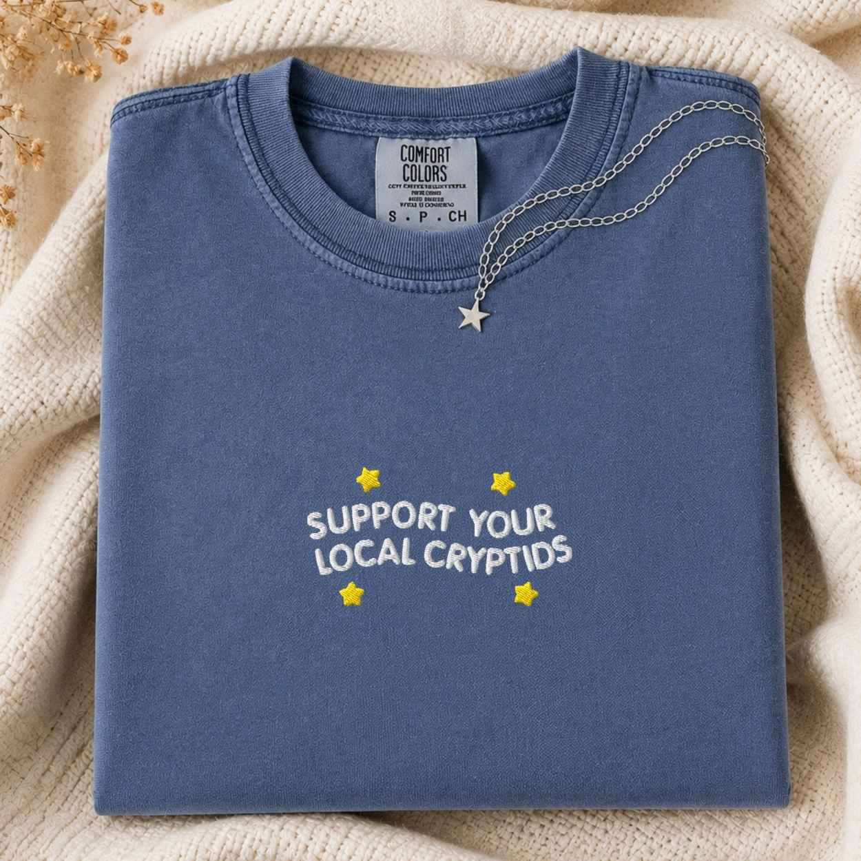 A folded slate blue t-shirt with 'SUPPORT YOUR LOCAL CRYPTIDS' and yellow stars printed on the front, styled on a cream knit blanket with a silver star necklace draped over the collar.
