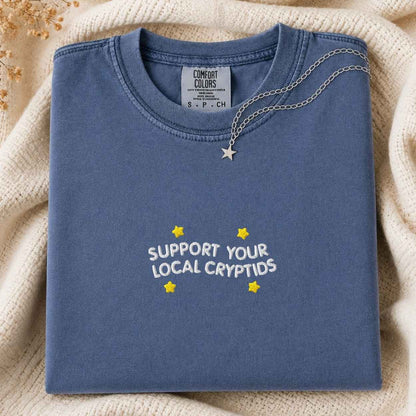A folded slate blue t-shirt with 'SUPPORT YOUR LOCAL CRYPTIDS' and yellow stars printed on the front, styled on a cream knit blanket with a silver star necklace draped over the collar.