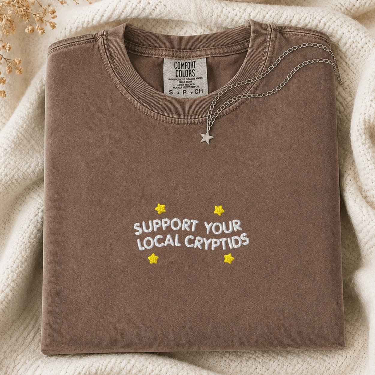 A folded brown sweatshirt with 'SUPPORT YOUR LOCAL CRYPTIDS' and yellow stars printed on the front, styled on a cream knit blanket with a silver star necklace. The neck label shows the brand Comfort Colors.