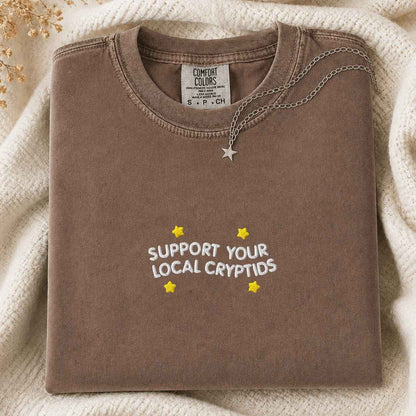 A folded brown sweatshirt with 'SUPPORT YOUR LOCAL CRYPTIDS' and yellow stars printed on the front, styled on a cream knit blanket with a silver star necklace. The neck label shows the brand Comfort Colors.