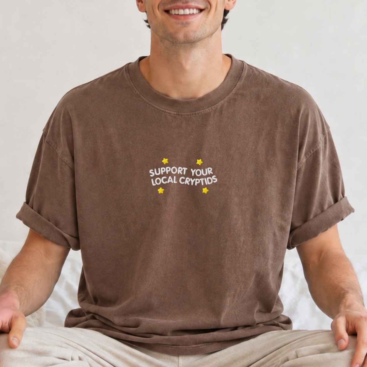 A man wearing a brown crewneck t-shirt with a text 'support your local cryptids'.