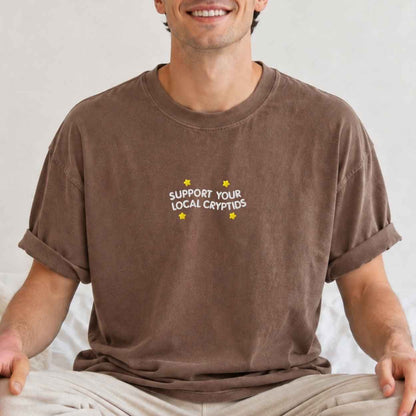 A man wearing a brown crewneck t-shirt with a text 'support your local cryptids'.