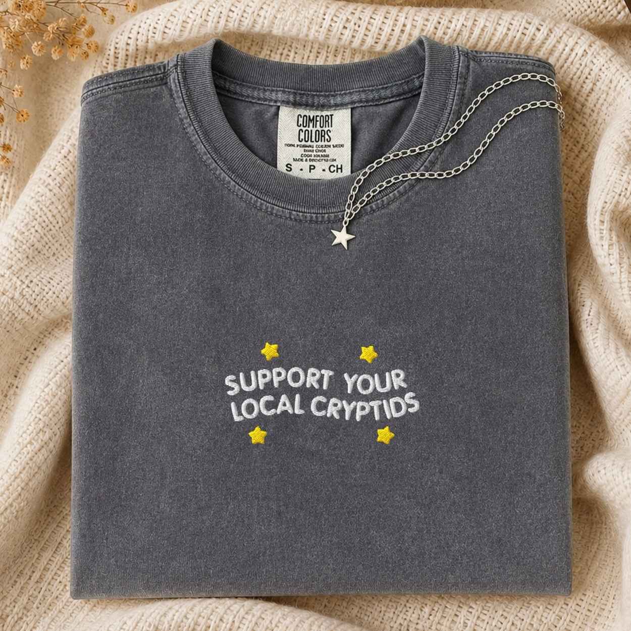 A folded dark gray t-shirt with 'SUPPORT YOUR LOCAL CRYPTIDS' printed in white and yellow, styled on a beige knit blanket with a silver necklace draped over the collar.