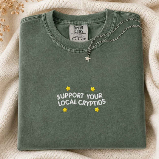 A folded sage green t-shirt with 'SUPPORT YOUR LOCAL CRYPTIDS' and yellow stars printed on the front, styled on a cream knit blanket with a silver star necklace.