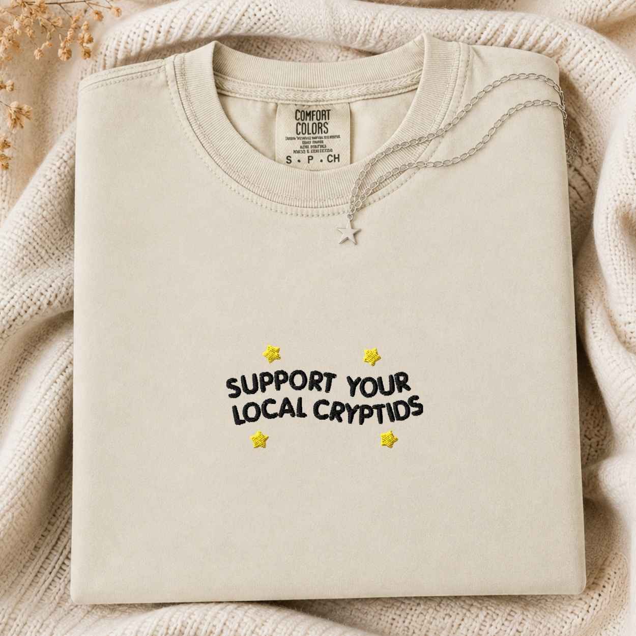 A folded cream-colored crewneck sweatshirt from Comfort Colors, featuring the text 'SUPPORT YOUR LOCAL CRYPTIDS' in black with yellow star accents, styled on a cozy cream knit blanket.