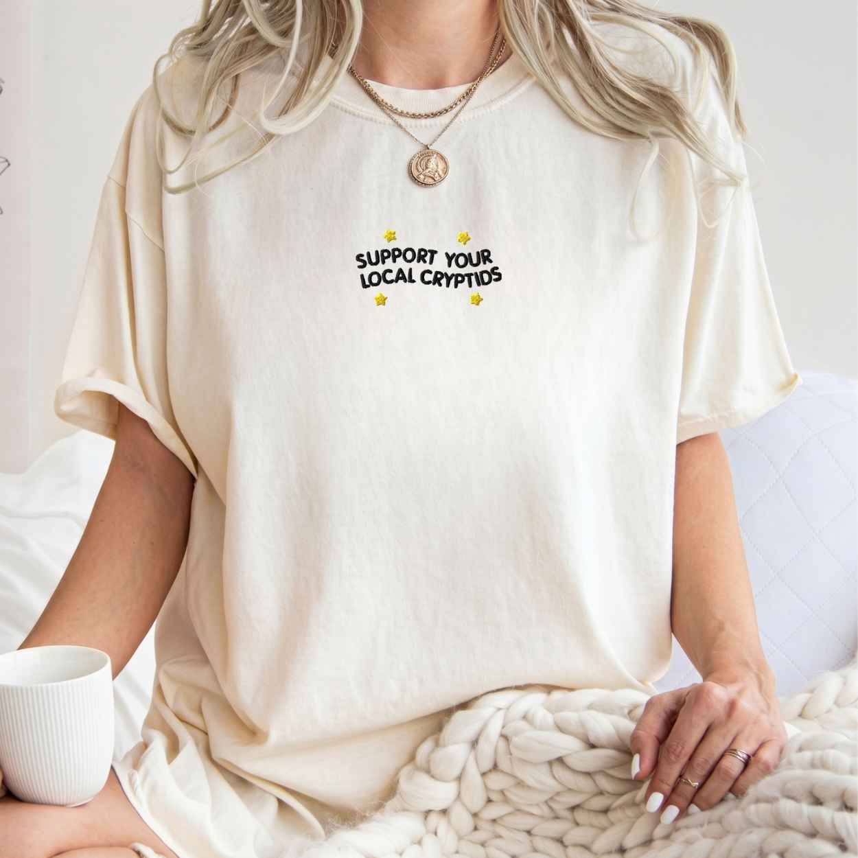 A woman wearing a cream-colored oversized t-shirt with the text 'SUPPORT YOUR LOCAL CRYPTIDS' printed in black on the chest, styled with a chunky knit blanket and a white mug.