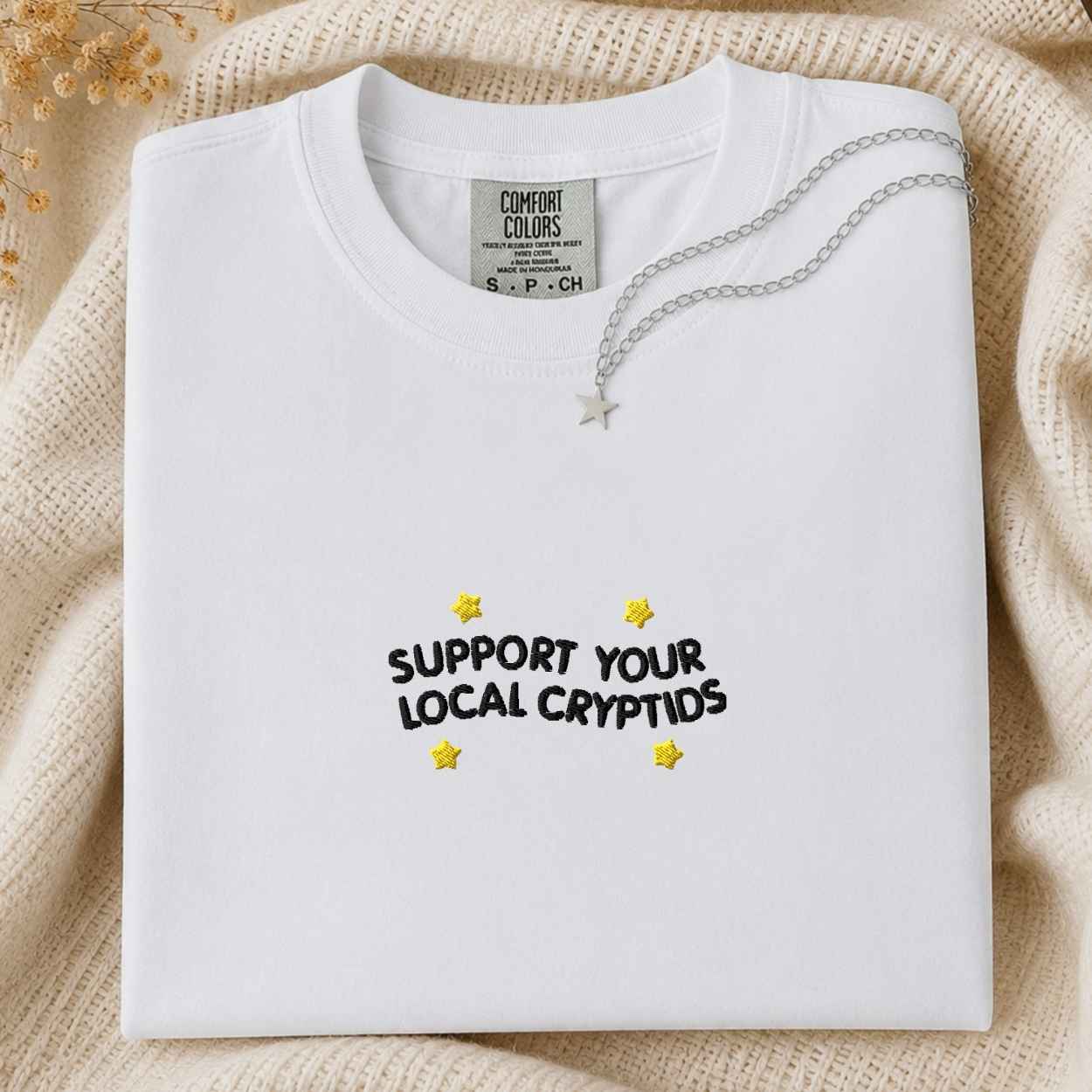A folded white t-shirt with 'SUPPORT YOUR LOCAL CRYPTIDS' written in black text and decorated with yellow stars, styled on a beige knit blanket with a silver necklace.