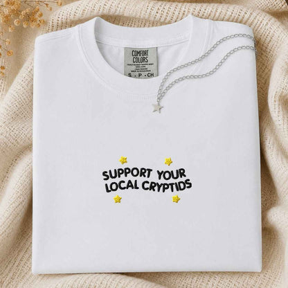 A folded white t-shirt with 'SUPPORT YOUR LOCAL CRYPTIDS' written in black text and decorated with yellow stars, styled on a beige knit blanket with a silver necklace.