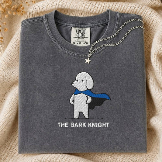 A folded dark gray Comfort Colors t-shirt featuring a white embroidered dog with a blue cape and the text 'THE BARK KNIGHT'. A silver star pendant necklace is draped over the collar.