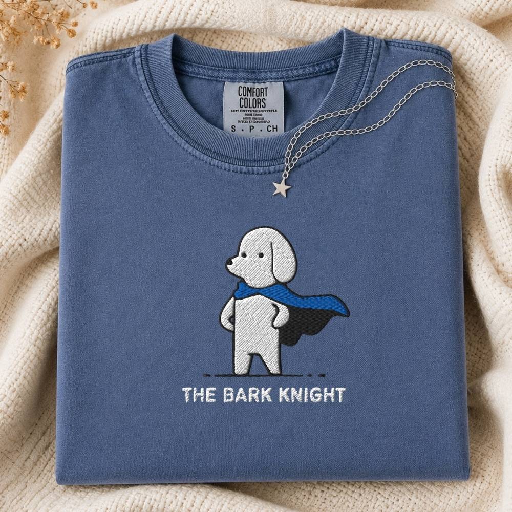 A folded slate blue crewneck tshirt with a graphic of a white dog in a black and blue cape and the text 'THE BARK KNIGHT' underneath. The sweatshirt is styled on a beige knit blanket with a silver necklace draped over the collar.
