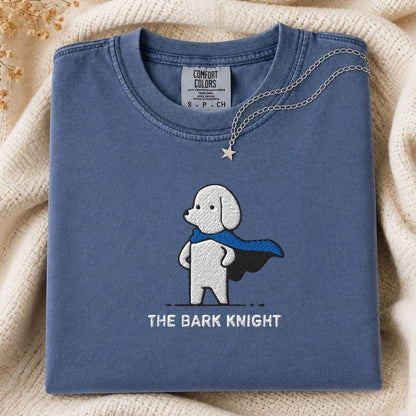 A folded slate blue crewneck tshirt with a graphic of a white dog in a black and blue cape and the text 'THE BARK KNIGHT' underneath. The sweatshirt is styled on a beige knit blanket with a silver necklace draped over the collar.