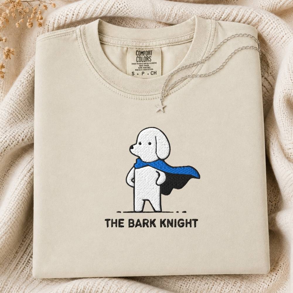 A folded cream-colored tee featuring a cartoon dog superhero graphic with a blue cape and the text 'THE BARK KNIGHT' underneath. The sweatshirt is displayed on a cozy, textured cream knit blanket with dried flowers in the corner.