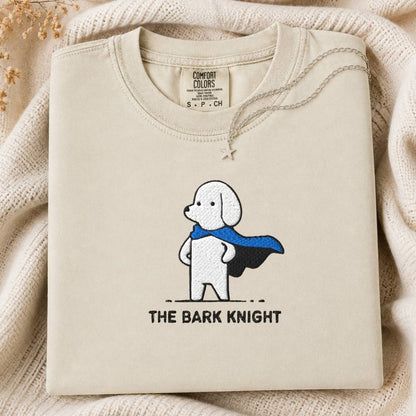 A folded cream-colored tee featuring a cartoon dog superhero graphic with a blue cape and the text 'THE BARK KNIGHT' underneath. The sweatshirt is displayed on a cozy, textured cream knit blanket with dried flowers in the corner.