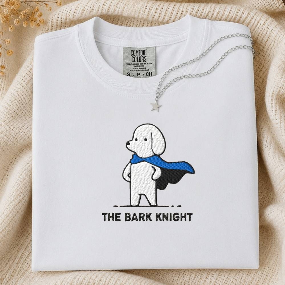 A folded white Comfort Colors T-shirt with a cartoon dog wearing a blue cape and the text 'THE BARK KNIGHT' printed below, styled on a beige knit blanket with a silver necklace draped across the collar.