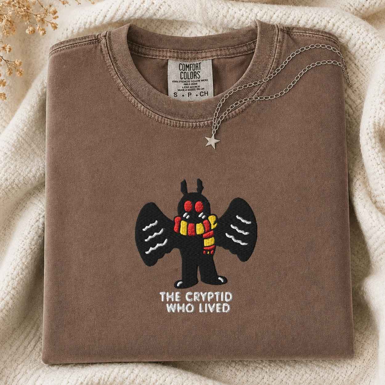 A folded brown tshirt with a graphic of a black mothman creature wearing a red and yellow striped scarf, with the text 'THE CRYPTID WHO LIVED' underneath. A silver star pendant necklace is draped over the collar.