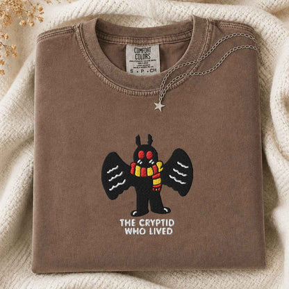 A folded brown tshirt with a graphic of a black mothman creature wearing a red and yellow striped scarf, with the text 'THE CRYPTID WHO LIVED' underneath. A silver star pendant necklace is draped over the collar.