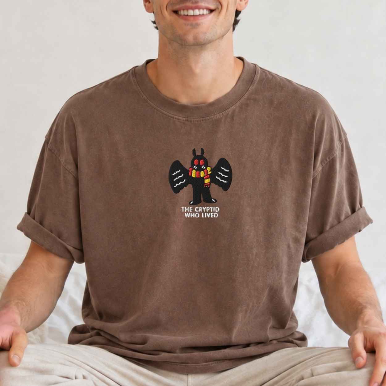 A man wearing a brown crewneck t-shirt with a graphic of a cartoon mothman and the text 'the cryptid who lived' on the chest, paired with light beige pants against a neutral background.