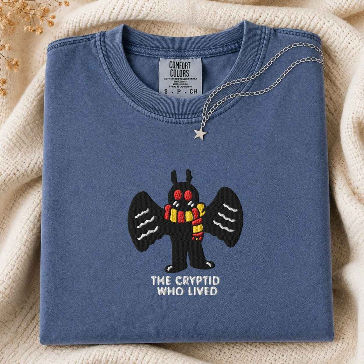 A folded slate blue t-shirt with a graphic of a black winged mothman creature wearing a red and yellow striped scarf, with the text 'THE CRYPTID WHO LIVED' underneath. A silver star necklace is draped over the collar.
