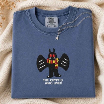A folded slate blue t-shirt with a graphic of a black winged mothman creature wearing a red and yellow striped scarf, with the text 'THE CRYPTID WHO LIVED' underneath. A silver star necklace is draped over the collar.