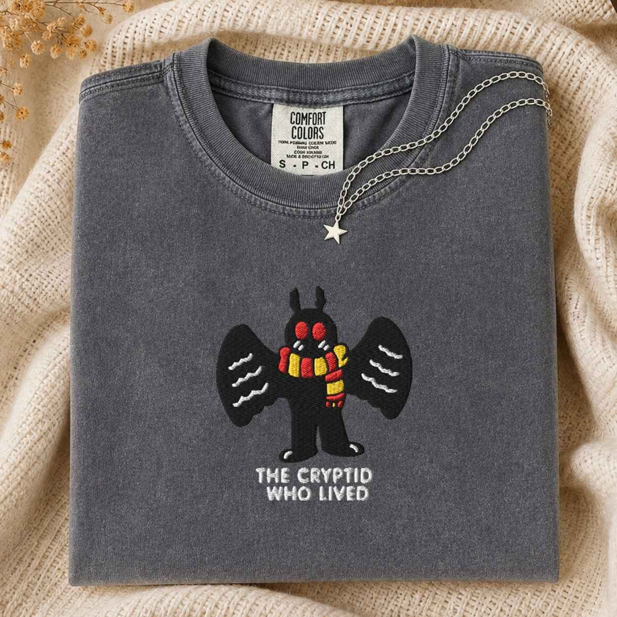 A folded dark gray t-shirt with a graphic embroidery of a black mothman cartoon creature wearing a red and yellow striped scarf, with the text 'THE CRYPTID WHO LIVED' underneath. A silver chain necklace with a star pendant is draped over the collar.