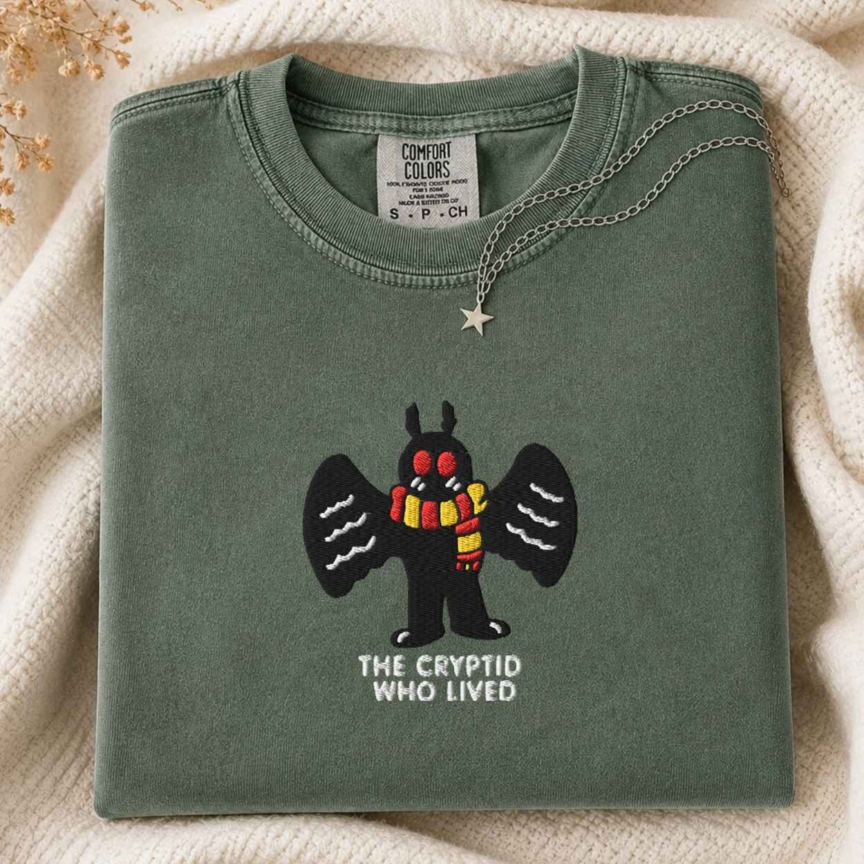 A folded sage green Comfort Colors t-shirt featuring a cartoon cryptid character with black wings and a red and yellow striped scarf, with the text 'THE CRYPTID WHO LIVED' underneath. The shirt is styled on a cream knit blanket with a silver star necklace draped over the collar.
