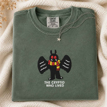 A folded sage green Comfort Colors t-shirt featuring a cartoon cryptid character with black wings and a red and yellow striped scarf, with the text 'THE CRYPTID WHO LIVED' underneath. The shirt is styled on a cream knit blanket with a silver star necklace draped over the collar.