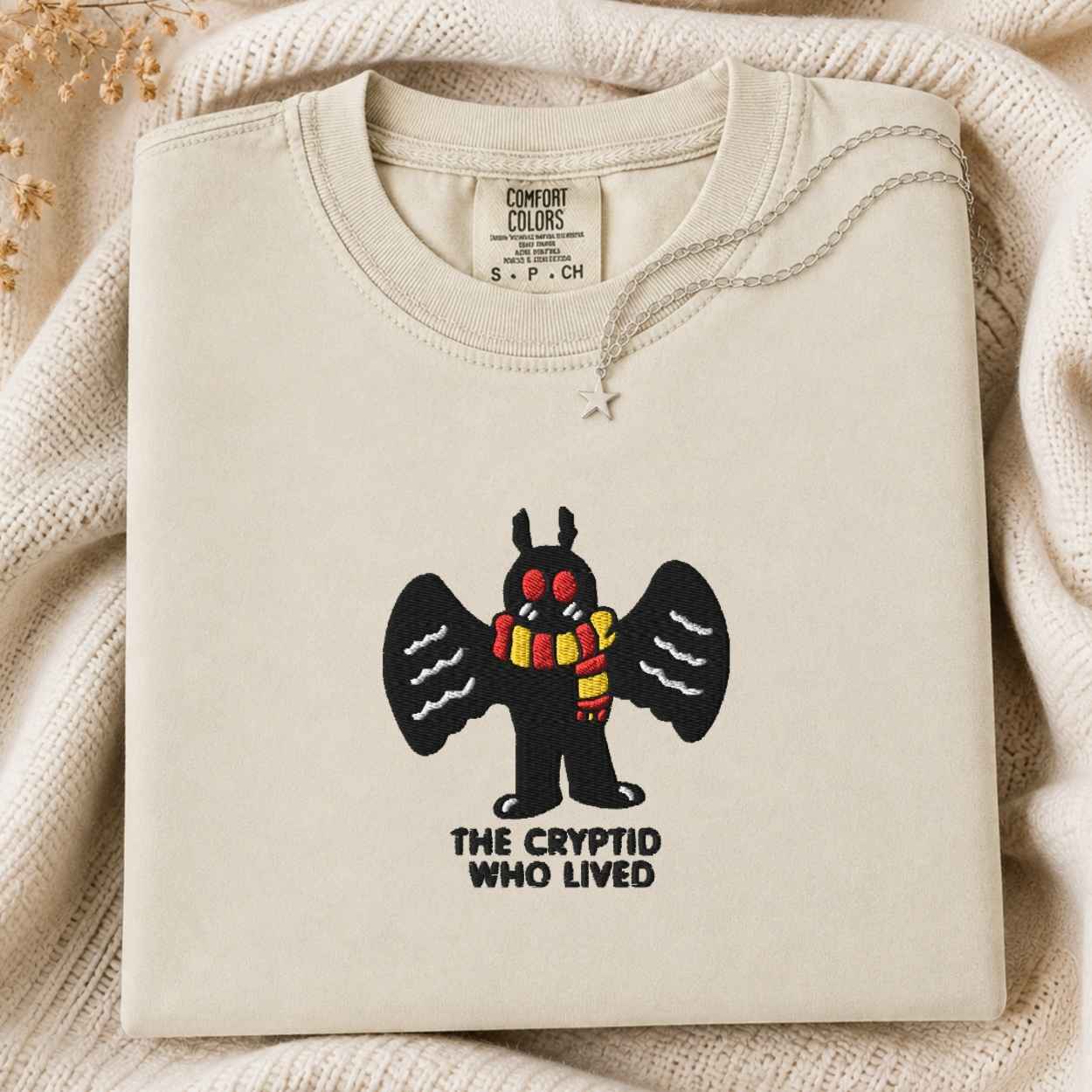 A folded cream-colored tshirt with a graphic of a black cartoon mothman cryptid with red eyes and a yellow-and-red striped scarf, above the text 'THE CRYPTID WHO LIVED'. The tshirt is styled on a textured cream knit blanket with dried flowers in the corner.
